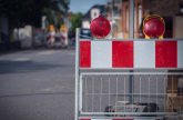 Straßensperrung Auf dem Bild sieht man eine Absperrung auf der Straße. Die Absperrung hat rote und weiße Streifen. Oben sind zwei rote Lampen, die blinken können. Im Hintergrund sieht man weitere Absperrungen und Häuser.