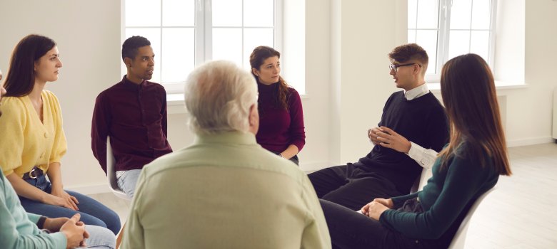 Gruppengespräch Das Bild zeigt eine Gruppe von sieben Personen, die in einem hellen Raum in einem Stuhlkreis sitzen. Die Menschen sind unterschiedlichen Alters und Geschlechts und blicken aufmerksam auf einen jungen Mann in schwarzem Pullover und Brille, der gerade spricht. Der Raum ist schlicht gestaltet, mit großen Fenstern, die viel Tageslicht hereinlassen.