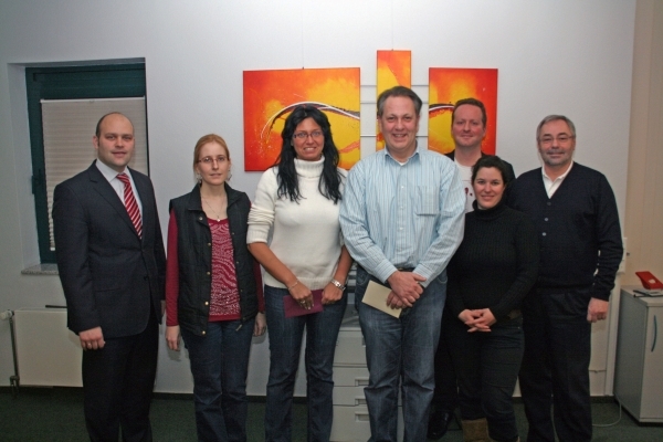 Das Bild zeigt sechs Personen, die nebeneinander vor einer weißen Wand stehen. Im Hintergrund hängen drei bunte Bilder mit orange-roten und gelben Farben. Alle Personen blicken freundlich in die Kamera. Die Gruppe besteht aus Männern und Frauen unterschiedlichen Alters. Links außen steht Bürgermeister Daniel Tybussek. 