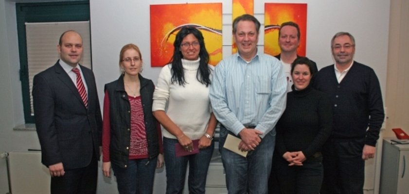 Das Bild zeigt sechs Personen, die nebeneinander vor einer weißen Wand stehen. Im Hintergrund hängen drei bunte Bilder mit orange-roten und gelben Farben. Alle Personen blicken freundlich in die Kamera. Die Gruppe besteht aus Männern und Frauen unterschiedlichen Alters. Links außen steht Bürgermeister Daniel Tybussek. 