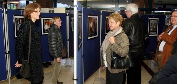 Das Bild zeigt mehrere Personen, die sich eine Ausstellung ansehen. Die Ausstellung besteht aus Fotografien, die auf blauen Stellwänden präsentiert werden. Die Besucherinnen und Besucher schauen sich die Bilder interessiert an. Der Raum hat große Fenster und wirkt hell und offen. Die Wände und der Boden sind in hellen Farben gestaltet.