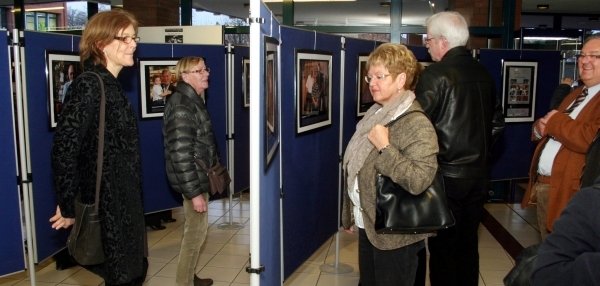 Das Bild zeigt mehrere Personen, die sich eine Ausstellung ansehen. Die Ausstellung besteht aus Fotografien, die auf blauen Stellwänden präsentiert werden. Die Besucherinnen und Besucher schauen sich die Bilder interessiert an. Der Raum hat große Fenster und wirkt hell und offen. Die Wände und der Boden sind in hellen Farben gestaltet.