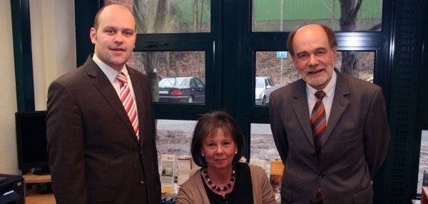 Das Bild zeigt zwei Männer und eine Frau in einem Büro. Die Frau sitzt an einem Schreibtisch und die beiden Männer stehen rechts und links neben ihr. Rechts von ihr steht Bürgermeister Daniel Tybussek. Alle drei lächeln in die Kamera. Im Hintergrund sind große Fenster mit Blick nach draußen zu sehen. Vor dem Fenster stehen Büroartikel und Informationsmaterial. Auf dem Schreibtisch liegen Dokumente, ein Taschenrechner und Büromaterialien.