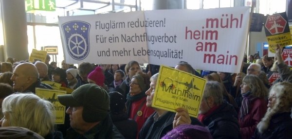 Das Bild zeigt viele Menschen auf einer Demonstration. Sie halten Schilder und Transparente hoch. Auf dem großen Banner steht: “Fluglärm reduzieren! Für ein Nachtflugverbot und mehr Lebensqualität!”. Daneben ist das Logo der Stadt Mühlheim am Main zu sehen. Viele der kleineren Schilder haben gelbe Hintergründe und fordern ebenfalls weniger Fluglärm. Die Menschen tragen warme Kleidung.