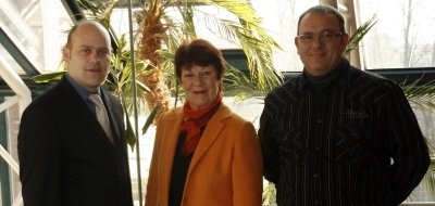 Das Bild zeigt drei Personen, die nebeneinander in einem hellen Raum stehen. Eine von ihnen ist Bürgermeister Daniel Tybussek. Im Hintergrund sind große Fenster und grüne Pflanzen zu sehen. Die Sonne scheint durch die Fenster und erzeugt eine freundliche Atmosphäre. Die Personen lächeln in die Kamera.