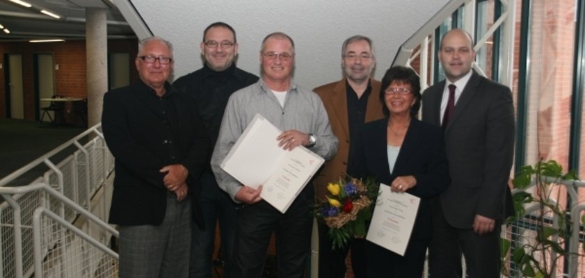 Das Bild zeigt sechs Personen, die zusammen auf einer Treppe stehen, darunter Bürgermeister Daniel Tybussek. Zwei Personen in der Mitte halten Urkunden in den Händen. Eine von ihnen hält zusätzlich einen Blumenstrauß. Alle lächeln freundlich in die Kamera. Der Raum hat große Fenster und viel Tageslicht.