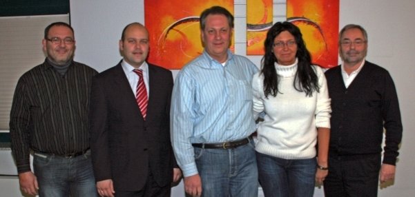 Das Bild zeigt fünf Personen, die nebeneinander vor einer weißen Wand stehen. In der Mitte hängen drei bunte Bilder mit orange-roten und gelben Farben. Die Personen sind drei Männer, darunter Bürgermeister Daniel Tybussek und eine Frau. Alle lächeln in die Kamera.