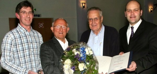 Das Bild zeigt vier Männer, die nebeneinander stehen. Der Mann ganz rechts ist Bürgermeister Daniel Tybussek. Er trägt einen schwarzen Anzug und eine Krawatte. Er hält eine geöffnete Urkunde in den Händen und lächelt. Der Mann neben ihm trägt eine Brille und eine dunkelblaue Jacke. Er hält einen großen Blumenstrauß mit weißen und blauen Blumen. Der dritte Mann von links trägt ein graues Jackett und eine Brille. Er lächelt freundlich in die Kamera. Der Mann ganz links trägt ein kariertes Hemd und hält ein kleines Geschenk in der Hand. Die Stimmung ist feierlich, und der Raum hat helle Wände mit dezenter Beleuchtung.