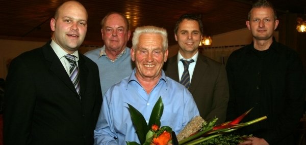 Das Bild zeigt fünf Männer, die zusammen in einem Raum stehen. Der Mann in der Mitte hat weißes Haar und trägt ein hellblaues Hemd. Er hält einen bunten Blumenstrauß in den Händen und lächelt in die Kamera. Die anderen vier Männer stehen links und rechts neben ihm. Zwei von ihnen tragen Anzüge und Krawatten, die anderen sind leger gekleidet. Der Raum hat eine Holzdecke und warme Beleuchtung.