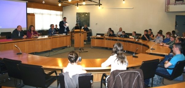 Das Bild zeigt einen großen Besprechungsraum mit vielen Menschen. Die Teilnehmenden sitzen an einem großen, halbkreisförmigen Holztisch. Vorne in der Mitte steht ein Rednerpult. Ein Mann spricht gerade zu den Anwesenden.  Links ist eine Leinwand mit einem blauen Bildschirm zu sehen. Die Decke des Raumes besteht aus Holz und hat viele Lampen.
