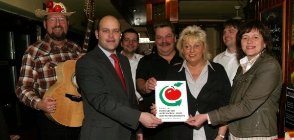 Das Bild zeigt eine Gruppe von sieben Personen in einem gemütlichen Raum. Im Mittelpunkt hält ein Mann im Anzug, Bürgermeister Daniel Tybussek, mit einer Frau ein Schild mit einem großen roten Apfel darauf. Auf dem Schild steht: "Förderung der hessischen und regionalen Obstwirtschaft". Links steht ein Mann mit einer Gitarre und einem Strohhut mit Deko-Äpfeln. Die anderen Personen lächeln ebenfalls freundlich in die Kamera. Der Raum hat warme Holzelemente und eine kleine Tafel im Hintergrund.