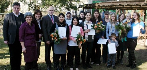 Das Bild zeigt eine große Gruppe von Menschen, die draußen im Grünen zusammenstehen. Viele Personen halten Blumensträuße und Urkunden in den Händen. Es sind sowohl Erwachsene als auch Kinder auf dem Bild zu sehen. Bürgermeister Daniel Tybussek steht ebenfalls dabei. Im Hintergrund sind Bäume, ein Holzhäuschen und ein Zaun zu erkennen.