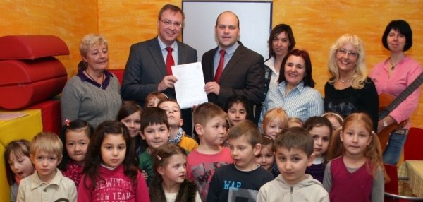Das Bild zeigt eine große Gruppe von Kindern und Erwachsenen in einem bunt gestalteten Raum. Im Vordergrund stehen viele Kinder unterschiedlichen Alters und schauen neugierig in die Kamera. Hinter den Kindern stehen mehrere Erwachsene. Zwei Männer in Anzügen, darunter Bürgermeister Daniel Tybussek, halten gemeinsam ein Dokument hoch und lächeln. Die Wände des Raumes sind in warmen Gelbtönen gestrichen. Links sind bunte Matten und Kissen zu sehen.