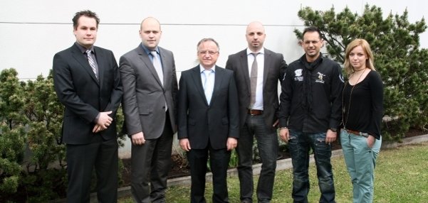 Das Bild zeigt sechs Personen, die draußen vor einer weißen Wand stehen. Hinter ihnen sind grüne Büsche zu sehen. Alle blicken freundlich in die Kamera. Die Gruppe besteht aus fünf Männern und einer Frau. Sie stehen auf einer gepflegten Rasenfläche.