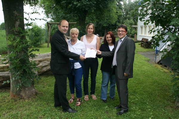 Das Bild zeigt fünf Personen, die draußen im Grünen stehen. In der Mitte halten alle gemeinsam ein Dokument. Sie lächeln freundlich in die Kamera. Der Hintergrund ist von Bäumen und Grasflächen geprägt. Rechts im Bild ist ein Gebäude mit weißen Fenstern zu sehen.