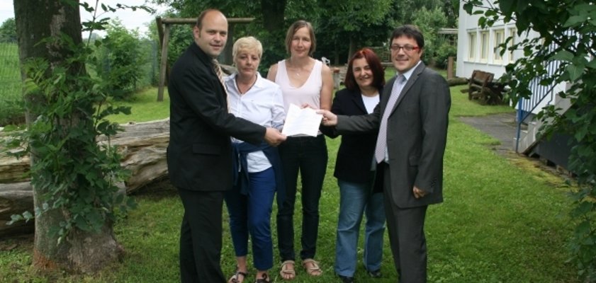Das Bild zeigt fünf Personen, die draußen im Grünen stehen. In der Mitte halten alle gemeinsam ein Dokument. Sie lächeln freundlich in die Kamera. Der Hintergrund ist von Bäumen und Grasflächen geprägt. Rechts im Bild ist ein Gebäude mit weißen Fenstern zu sehen.