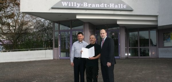 Das Bild zeigt drei Männer, die auf einem gepflasterten Platz vor einer Halle stehen. Über dem Eingang der Halle ist ein großes Schild mit der Aufschrift "Willy-Brand-Halle" zu sehen. Einer der Männer ist Bürgermeister Daniel Tybussek. Der Mann neben ihm hält eine Urkunde in den Händen, die sie gemeinsam präsentieren. Alle lächeln freundlich in die Kamera. Im Hintergrund sind Glasfenster und einige Bäume zu erkennen.