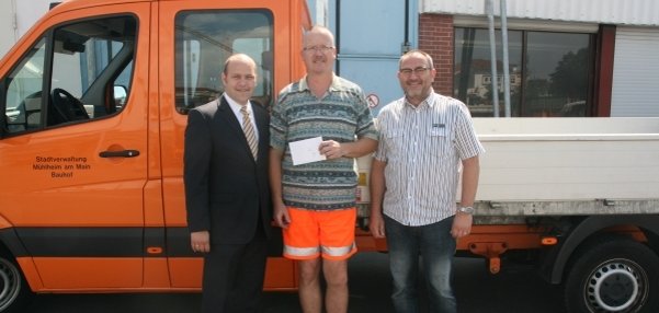 Das Bild zeigt drei Männer, die vor einem orangefarbenen Fahrzeug stehen. Das Fahrzeug gehört zur Stadtverwaltung Mühlheim am Main, Bereich Bauhof. Es hat eine leuchtende orange Farbe und eine Ladefläche. Der Mann in der Mitte trägt Arbeitskleidung mit einer leuchtend orangefarbenen Shorts und häkt ein weißes Kuvert in der Hand. Die beiden Männer links und rechts von ihm, darunter Bürgermeister Daniel Tybussek, lächeln ebenfalls in die Kamera. Im Hintergrund sind ein Gebäude mit blauen und weißen Wänden sowie eine offene Garage zu sehen.