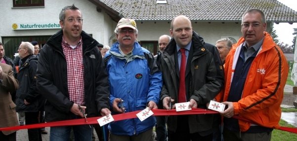 Das Bild zeigt vier Männer, die ein rotes Band durchschneiden. Jeder hält eine Schere in der Hand und gemeinsam schneiden sie das Band an mehreren Stellen durch. Im Hintergrund steht ein weißes Gebäude mit der Aufschrift "Naturfreundehaus". Weitere Personen schauen dem Banddurchschnitt zu.