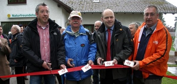 Das Bild zeigt vier Männer, die ein rotes Band durchschneiden. Jeder hält eine Schere in der Hand und gemeinsam schneiden sie das Band an mehreren Stellen durch. Im Hintergrund steht ein weißes Gebäude mit der Aufschrift "Naturfreundehaus". Weitere Personen schauen dem Banddurchschnitt zu.