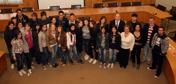 Das Bild zeigt eine große Gruppe junger Menschen in einem Besprechungsraum. Die Gruppe besteht aus Jugendlichen und einigen Erwachsenen. Sie stehen eng beieinander und blicken lächelnd in die Kamera. Im Hintergrund ist ein großer Konferenztisch aus Holz zu sehen, sowie eine Leinwand an der Wand. Die Decke hat schwarze Lampen. Durch die Fenster scheint Tageslicht.