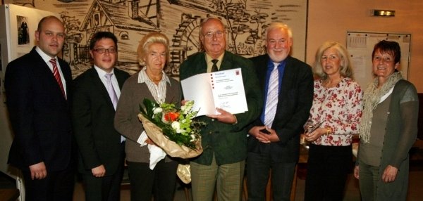 Das Bild zeigt sieben Personen, die gemeinsam in einem Raum stehen. Der Mann in der Mitte hält eine große Urkunde in den Händen. Er lächelt in die Kamera. Neben ihm steht eine Frau mit einem Blumenstrauß. Die anderen Personen blicken ebenfalls freundlich in die Kamera. Im Hintergrund ist eine große Wanddekoration mit einem historischen Motiv zu sehen. Auf dem Tisch vorne liegt eine goldene Tischdecke mit einigen Papieren.