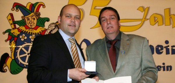 Das Bild zeigt zwei Männer, darunter Bürgermeister Daniel Tybussek. Sie stehen vor einem festlichen Hintergrund mit einer großen Zahl "55" und bunten Karnevalsmotiven.  Links ist ein bunt gekleideter Narr mit einem Rad als Symbol zu sehen. Der Bürgermeister steht links im Bild. Er hält eine kleine Schatulle mit einer Medaille in der Hand. Der Mann rechts neben ihm präsentiert eine geöffnete Urkunde. Beide Männer lächeln in die Kamera.