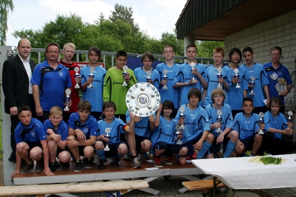 Das Bild zeigt eine Jugendfußballmannschaft in hellblauen Trikots. Alle Spieler und Trainer halten Pokale in den Händen. In der Mitte wird eine große Meisterschale präsentiert. Die Mannschaft steht auf einer kleinen Bühne im Freien. Im Hintergrund sind grüne Bäume und ein Gebäude mit heller Steinwand zu sehen. Bürgermeister Daniel Tybussek steht links im Bild. Im Vordergrund stehen Biertische und Bänke.