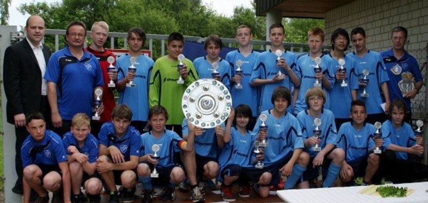 Das Bild zeigt eine Jugendfußballmannschaft in hellblauen Trikots. Alle Spieler und Trainer halten Pokale in den Händen. In der Mitte wird eine große Meisterschale präsentiert. Die Mannschaft steht auf einer kleinen Bühne im Freien. Im Hintergrund sind grüne Bäume und ein Gebäude mit heller Steinwand zu sehen. Bürgermeister Daniel Tybussek steht links im Bild. Im Vordergrund stehen Biertische und Bänke.