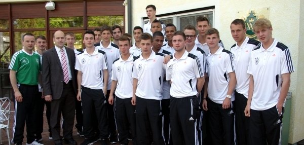 Das Bild zeigt eine Gruppe junger Fußballspieler in einheitlicher Sportkleidung. Alle Spieler tragen weiße Trainingsshirts mit schwarzen Streifen und schwarze Trainingshosen. Sie stehen eng beieinander und schauen freundlich in die Kamera. Links im Bild steht ein Mann im grünen Traineroutfit und Bürgermeister Daniel Tybussek im Anzug. Der Hintergrund zeigt ein Gebäude mit hellen Wänden und großen Fenstern. Auch ein roter Pflasterboden ist zu sehen.