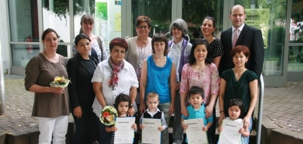 Das Bild zeigt eine Gruppe von Erwachsenen und Kindern. Sie stehen vor einem Gebäude mit Glas-Eingangstüren. Die Erwachsenen stehen im Hintergrund. Die Kinder stehen vorne und halten Urkunden in den Händen. Einige Personen haben kleine Blumensträuße dabei. Alle blicken freundlich in die Kamera. Der Platz ist gepflastert und von Grünpflanzen umgeben.