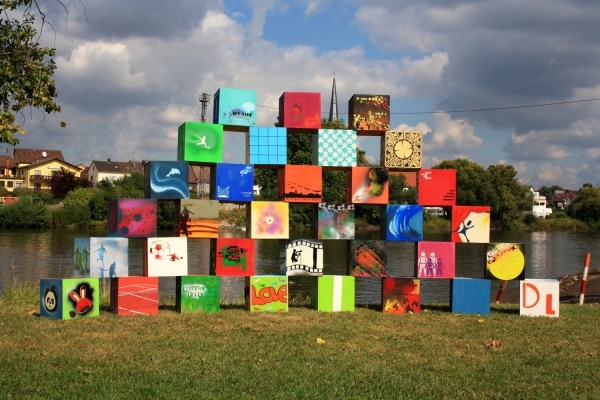 Das Bild zeigt eine bunte Installation aus vielen quadratischen Boxen. Sie sind zu einer Pyramide aufgestapelt. Jede Box ist anders bemalt. Mit bunten Farben, Mustern, Symbolen und Bildern. Man sieht Motive wie eine Uhr, das Wort "LOVE", Sportfiguren und abstrakte Kunst. Die Installation seht auf einer grünen Wiese direkt am Wasser. Im Hintergrund erkennt man Bäume, Häuser und einen Kirchturm. Der Himmel ist blau mit weißen Wolken.