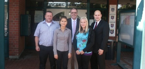 Das Bild zeigt fünf Personen, die vor dem Eingang des Rathauses stehen. Über ihnen hängt ein großes Schild mit der Aufschrift "Eingang Rathaus". Die Gruppe besteht aus drei Männern und zwei Frauen. Alle stehen dicht beieinander und lächeln freundlich in die Kamera. Die Umgebung ist geprägt von roten Klinkersteinen, Glasfenstern und Informationstafeln rund um den Rathauseingang.