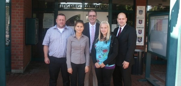 Das Bild zeigt fünf Personen, die vor dem Eingang des Rathauses stehen. Über ihnen hängt ein großes Schild mit der Aufschrift "Eingang Rathaus". Die Gruppe besteht aus drei Männern und zwei Frauen. Alle stehen dicht beieinander und lächeln freundlich in die Kamera. Die Umgebung ist geprägt von roten Klinkersteinen, Glasfenstern und Informationstafeln rund um den Rathauseingang.