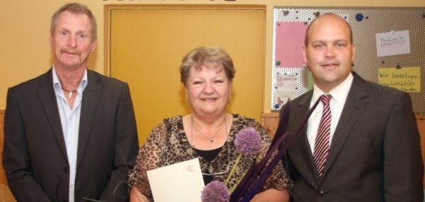 Das Bild zeigt drei Personen, die vor einer gelben Tür stehen. Die Frau in der Mitte hält einen großen, bunten Blumenstrauß und eine weiße Urkunde in den Händen. Sie lächelt freundlich in die Kamera. Links steht ein Mann im Anzug und rechts Bürgermeister Daniel Tybussek. Er trägt ebenfalls einen Anzug. Im Hintergrund sind bunte Pinnwände mit Notizen zu sehen.