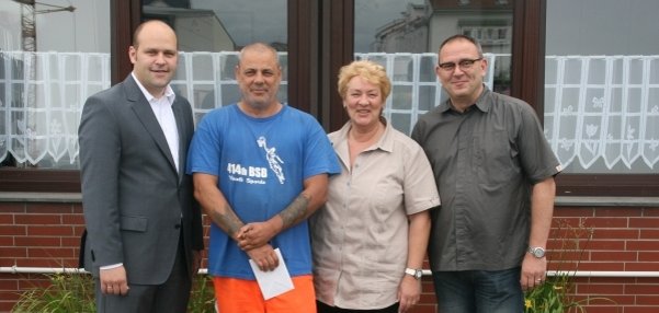 Das Bild zeigt vier Personen, die vor einem Gebäude mit roten Klinkerwänden und großen Fenstern stehen. Der Mann in der Mitte trägt Arbeitskleidung mit einem blauen T-Shirt und leichtend orangefarbenen Hosen. Er hält ein weißes Kuvert in den Händen. Links steht Bürgermeister Daniel Tybussek im Anzug. Rechts steht eine Frau in beiger Kleidung und  ein weiterer Mann in Freizeitkleidung. Alle lächeln freundlich in die Kamera. Im Hintergrund sind weiße Blumenkästen mit grünen Pflanzen zu sehen.