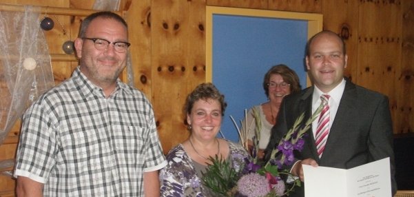 Das Bild zeigt drei Personen, die lächelnd in die Kamera blicken. Die Frau in der Mitte hält einen großen Blumenstrauß mit lila Blumen in den Händen. Der Mann rechts im Anzug, Bürgermeister Daniel Tybussek, präsentiert eine geöffnete Urkunde. Links steht ein weiterer Mann in einem karierten Hemd. Im Hintergrund ist eine weitere Frau zu sehen. Die Wände sind aus hellem Holz. Auf dem Tisch links stehen Gläser und Dekorationen.