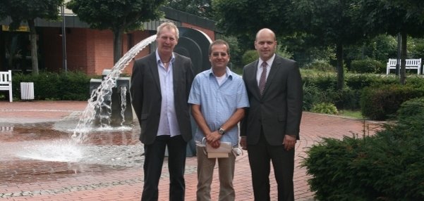 Das Bild zeigt drei Männer, die auf einem gepflasterten Platz vor einem Springbrunnen stehen, Der Brunnen im Hintergrund sprudelt Wasser in die Luft. Rundherum sind grüne Büsche, Bäume und weiße Bänke zu sehen. Die Männer lächeln in die Kamera. Der Mann in der Mitte hält ein Dokument in den Händen.