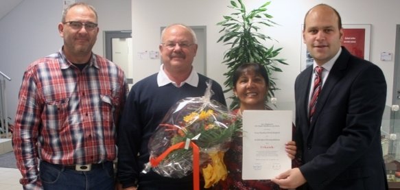 Das Bild zeigt vier Personen, die in einem hellen Raum stehen. Eine Frau in der Mitte hält einen großen, bunten Blumenstrauß und eine Urkunde in den Händen. Bürgermeister Daniel Tybussek steht rechts neben ihr. Er trägt einen Anzug und unterstützt sie beim Halten der Urkunde. Die beiden anderen Männer stehen links und lächeln ebenfalls in die Kamera. Im Hintergrund ist eine grüne Pflanze und eine Vitrine zu sehen.