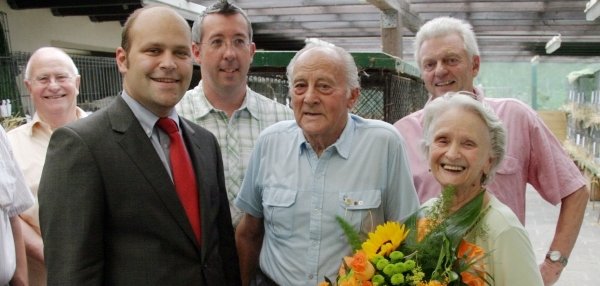 Das Bild zeigt sechs Personen, die in einem offenen Raum stehen. Sie lächeln freundlich in die Kamera. Eine ältere Frau hält einen großen, bunten Blumenstrauß mit gelben und orangenen Blumen in den Händen. Neben ihr steht ein älterer Mann in einem hellblauen Hemd. Links im Bild steht Bürgermeister Daniel Tybussek im Anzug. Im Hintergrund stehen weitere Personen.