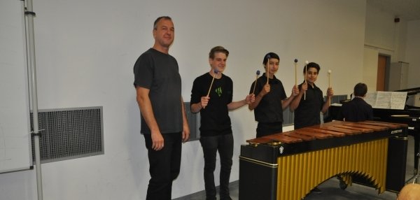 Das Bild zeigt vier Männer in einem Musikraum. Drei der Männer stehen hinter einem großen Marimbaphon. Sie halten Schläger in den Händen und lächeln in die Kamera. Der vierte Mann steht links neben dem Instrument. Im Hintergrund ist ein Klavier zu sehen. An dem Klavier sitzt eine weitere Person. Die Wände des Raums sind schlicht weiß.