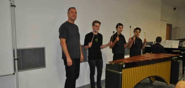 Das Bild zeigt vier Männer in einem Musikraum. Drei der Männer stehen hinter einem großen Marimbaphon. Sie halten Schläger in den Händen und lächeln in die Kamera. Der vierte Mann steht links neben dem Instrument. Im Hintergrund ist ein Klavier zu sehen. An dem Klavier sitzt eine weitere Person. Die Wände des Raums sind schlicht weiß.