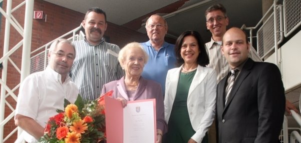 Schöffin des Ortsgerichtes Mühlheim II Das Bild zeigt eine Gruppe von sieben Personen, die auf einer Treppe stehen. In der Mitte hält eine Frau eine geöffnete Urkunde in den Händen. Sie lächelt freundlich in die Kamera. Links von ihr steht ein Mann mit einem bunten Blumenstrauß. Die anderen Personen umrahmen die Szene und wirken ebenfalls fröhlich. Unter ihnen ist Bürgermeister Daniel Tybussek. Der Hintergrund zeigt eine helle Treppenanlage mit Metallgeländern und Backsteinwänden.