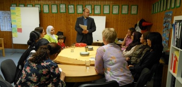Das Bild zeigt eine Gruppe von Menschen. Sie sitzen an einem großen runden Tisch. Ein Mann im Anzug, Bürgermeister Daniel Tybussek, steht vor der Holzvertäfelten Wand. Er spricht zu den Anwesenden. Er hält ein Dokument in den Händen. Die anderen Personen hören aufmerksam zu, Einige tragen bunte Kleidung und Kopftücher. Im Hintergrund sind eine Tafel, bunte Plakate und Bücherregale zu sehen.