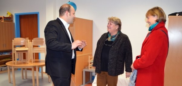 Das Bild zeigt drei Personen, die sich in einem hellen Raum unterhalten. Links steht Bürgermeister Daniel Tybussek im schwarzen Anzug. Ihm gegenüber stehen zwei Frauen, die ihm aufmerksam zuhören. Die Frau in der Mitte trägt eine dunkle Jacke, die Frau rechts einen auffälligen roten Mantel. Im Hintergrund stehen gestapelte Holzstühle auf einem runden Tisch. An der Wand sind helle Schränke und ein blau umrahmter Türrahmen zu sehen. Die Atmosphäre wirkt freundlich und interessiert.