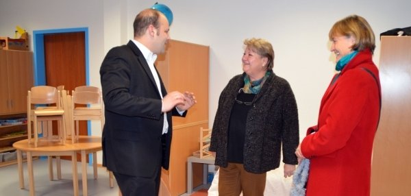 Das Bild zeigt drei Personen, die sich in einem hellen Raum unterhalten. Links steht Bürgermeister Daniel Tybussek im schwarzen Anzug. Ihm gegenüber stehen zwei Frauen, die ihm aufmerksam zuhören. Die Frau in der Mitte trägt eine dunkle Jacke, die Frau rechts einen auffälligen roten Mantel. Im Hintergrund stehen gestapelte Holzstühle auf einem runden Tisch. An der Wand sind helle Schränke und ein blau umrahmter Türrahmen zu sehen. Die Atmosphäre wirkt freundlich und interessiert.