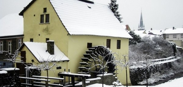 Auf dem Bild ist die Brückenmühle von Mühlheim zu sehen. Die Mühle ist ein gelbes Haus mit einem großen Wasserrad an der Seite. Alles ist mit Schnee bedeckt. Im Hintergrund sind weitere Häuser und ein Kirchturm zu sehen. Vor der Mühle stehen kleine, kahle Bäume. Die Szene wirkt ruhig und idyllisch.