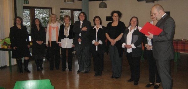 Mehrere Frauen stehen in einer Reihe in einem Raum mit Holzboden. Die Frauen tragen überwiegend schwarze Hosen und weiße Blusen oder Hemden. Rechts im Bild steht Bürgermeister Daniel Tybussek mit einem Ordner in der Hand. Er liest aus dem Ordner vor. Im Hintergrund sind große Fenster mit Blick auf schneebedeckte Bäume zu sehen. Alle Personen wirken aufmerksam und konzentriert.
