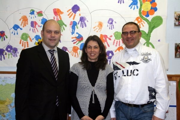 Drei Personen stehen nebeneinander und lächeln in die Kamera. Hinter ihnen ist eine bunte Wand mit vielen Handabdrücken in verschiedenen Farben. Links steht Bürgermeister Daniel Tybussek im Anzug. In der Mitte eine Frau mit dunklen Haaren. Rechts steht ein Mann mit Brille und weißem Hemd. Die Atmosphäre wirkt freundlich und bunt.