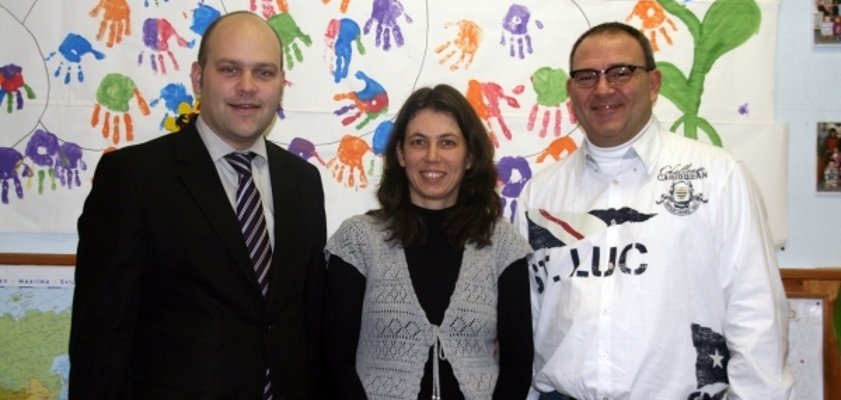 Drei Personen stehen nebeneinander und lächeln in die Kamera. Hinter ihnen ist eine bunte Wand mit vielen Handabdrücken in verschiedenen Farben. Links steht Bürgermeister Daniel Tybussek im Anzug. In der Mitte eine Frau mit dunklen Haaren. Rechts steht ein Mann mit Brille und weißem Hemd. Die Atmosphäre wirkt freundlich und bunt.